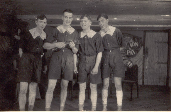 Four men dressed in rompers sing to audience, one playing a ukelele-like instument