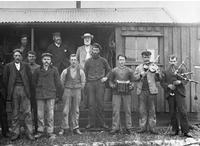 Photo of men  (including a bagpiper) in front of simple house 